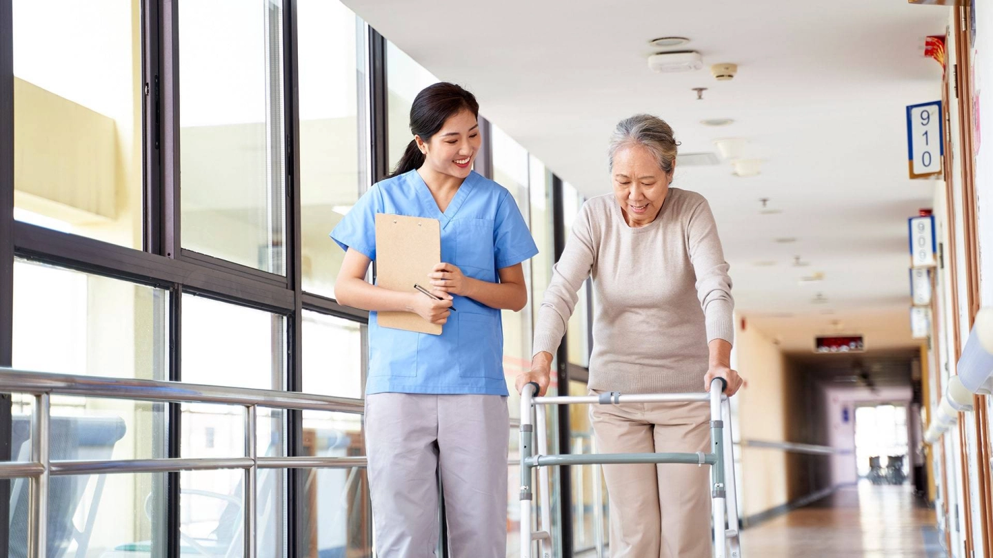 Сиделка сопровождает пожилую женщину, которая передвигается с помощью ходунков.