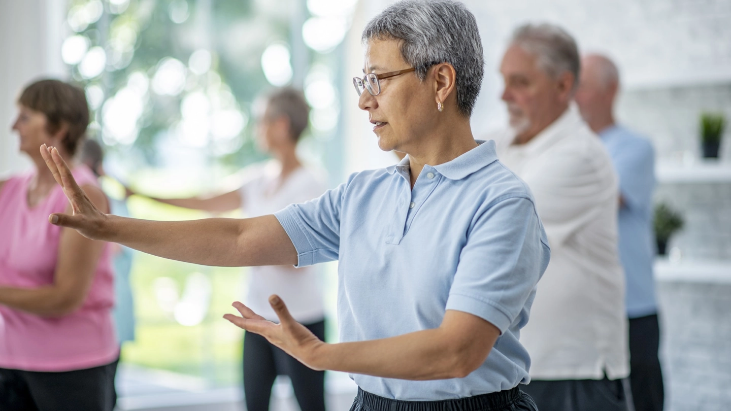 Abwehrkräfte: Mehrere Menschen stehen in einem hellen Raum und führen Bewegungsübungen durch. Im Vordergrund steht eine ältere Frau die mit beiden Armen eine Bewegung ausführt.