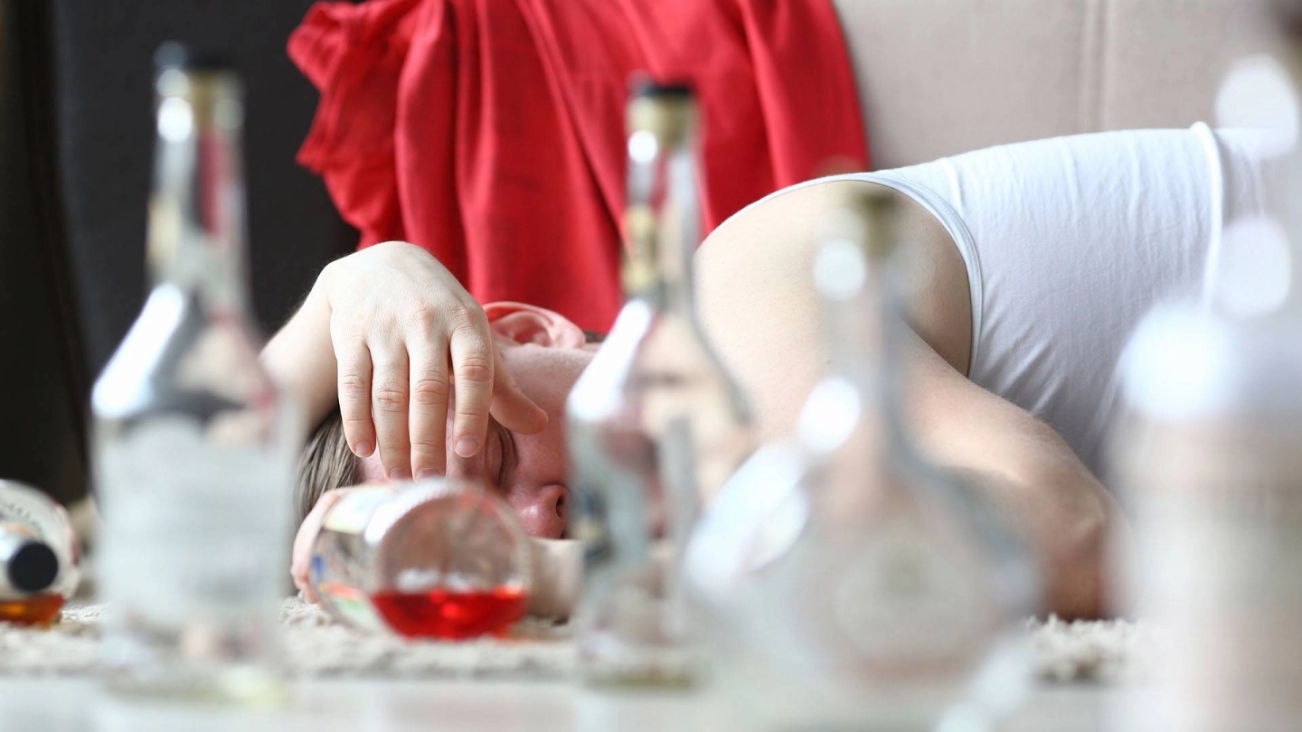 Auf einem Tisch sind verschwommen Flaschen und Gläser zu sehen. Eine Person liegt daneben und schläft.