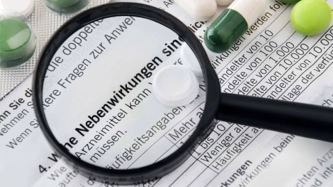 Part of a package insert with several tablets lying on it. The sentence “What side effects are possible?” is enlarged by a magnifying glass.