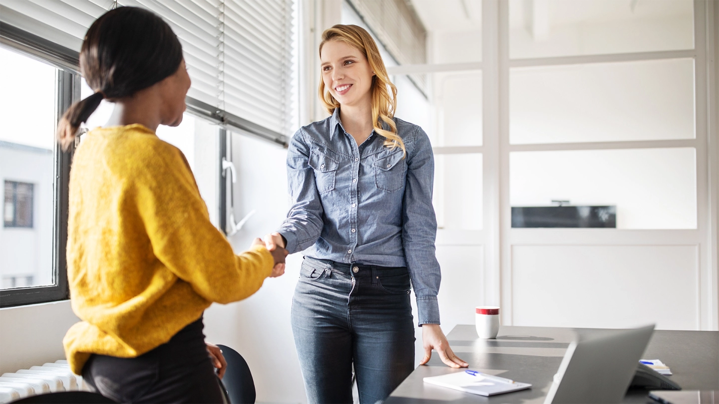 Betriebliches Eingliederungsmanagement: Zwei Frauen stehen an einem Bürotisch und schütteln sich nach einem erfolgreichen geschäftlichen Gespräch zustimmend die Hände.