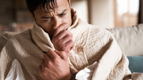 Entzündung der Atemwege: Ein Mann sitzt auf einer Couch, hat seinen kompletten Oberkörper in eine wärmende Decke gehüllt und greift sich mit einer Hand an die Nase. Sie scheint verschnupft zu sein.