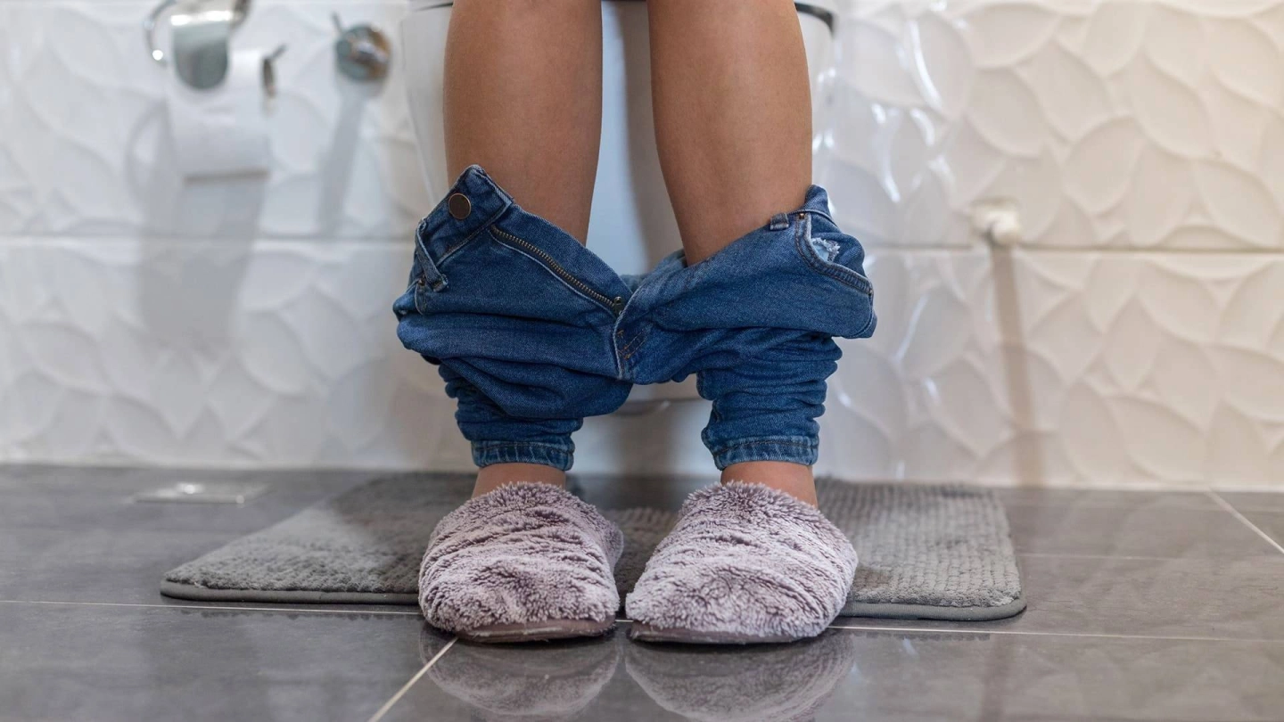 Eine Person sitzt mit heruntergelassenen Hosen und Hausschuhen an den Füßen auf der Toilette.