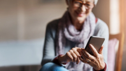 Пожилая женщина в очках сидит на диване. Женщина держит в левой руке смартфон, правой рукой перемещается по экрану и улыбается.