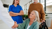 Eine ältere Frau sitzt in einem Rollstuhl in einem hellen Krankenhausflur. Sie lächelt und gibt einer Ärztin oder einem Arzt die Hand. Im Hintergrund stehen eine Krankenpflegerin und ein älterer Mann, der den Rollstuhl schiebt und eine Tasche trägt.