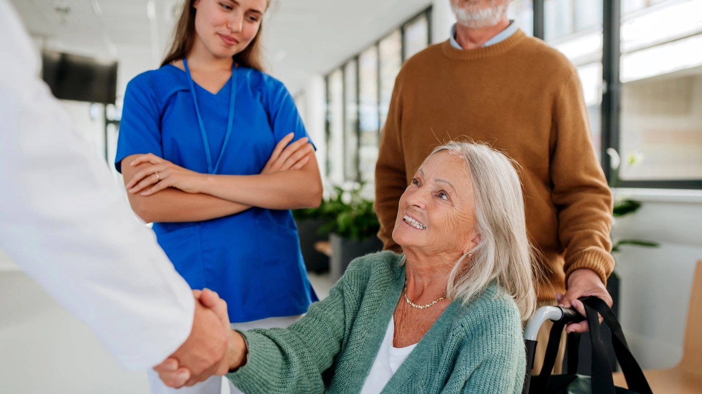 Eine ältere Frau sitzt in einem Rollstuhl in einem hellen Krankenhausflur. Sie lächelt und gibt einer Ärztin oder einem Arzt die Hand. Im Hintergrund stehen eine Krankenpflegerin und ein älterer Mann, der den Rollstuhl schiebt und eine Tasche trägt.