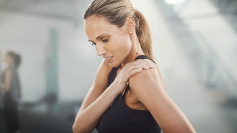 Eine Frau in Sportkleidung und leicht schmerzverzerrtem Blick greift sich an die Schulter.