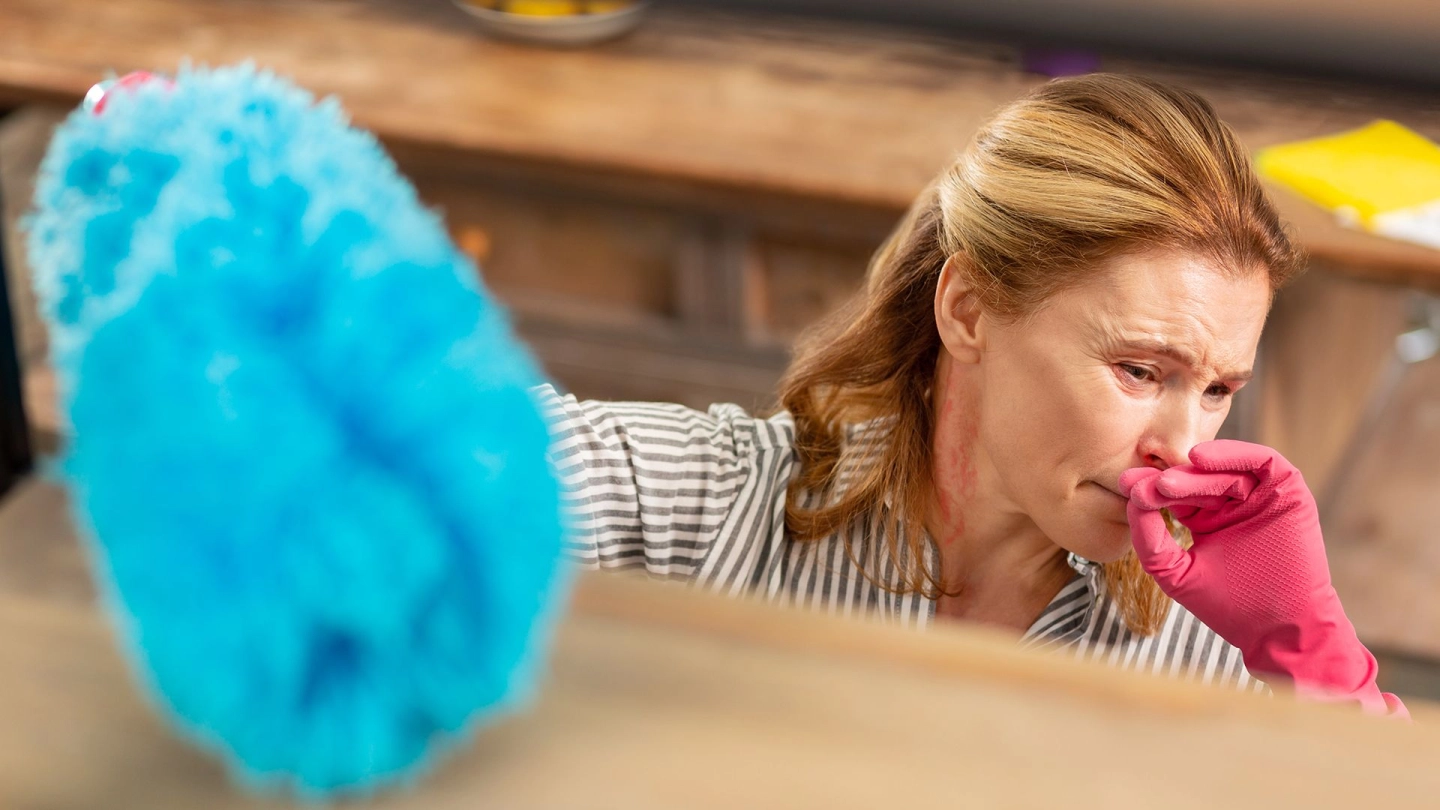 Аллергия на домашнюю пыль: женщина стирает пыль и касается лица рукой. Ее глаза покраснели.