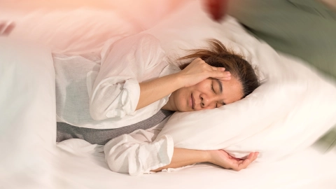 Женщина лежит в постели, схватившись правой рукой за висок. Ее глаза закрыты, а лицо напряжено.