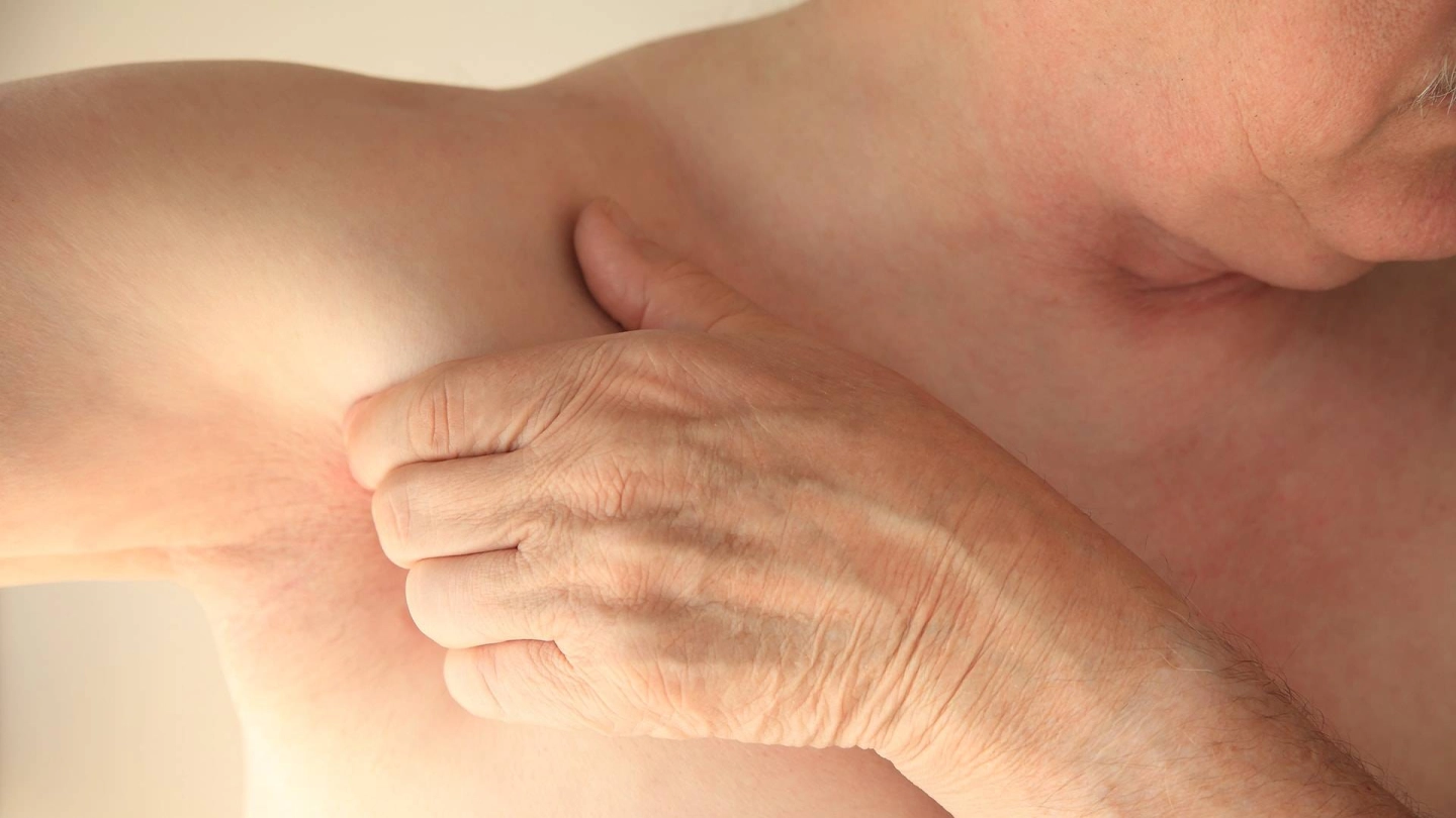 Ein Mann mit freiem Oberkörper streckt seinen rechten Arm zur rechten Seite aus. Mit der linken Hand umgreift er seine Achselhöhle.