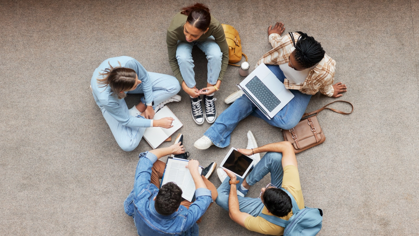 Junge Studierende mit Arbeitsmaterialien sitzen auf dem Fußboden im Kreis.