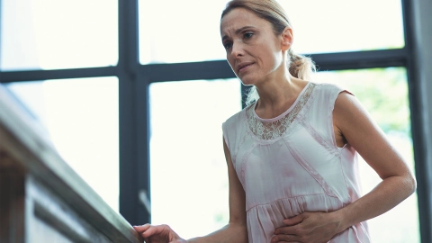 Magengeschwür: Eine Frau steht in ihrer Wohnung und hält sich den Bauch. Sie wirkt angespannt und als hätte sie Schmerzen.