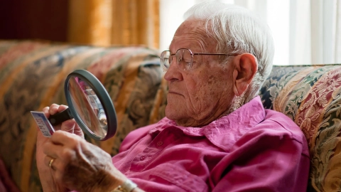 Ein älterer Mann sitzt auf einem Sessel. Er hält eine große Lupe in der Hand und blickt durch sie auf eine Plastikkarte.
