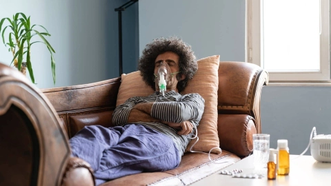 Ein Mann liegt auf dem Sofa. Er ist über eine Maske an ein Beatmungsgerät angeschlossen. Auf dem Tisch neben ihm stehen Medikamente.