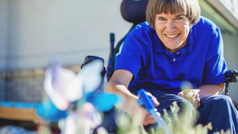 Eine Frau mittleren Alters sitzt im Rollstuhl und pflegt ihre Blumen mit einem Handgreifer.