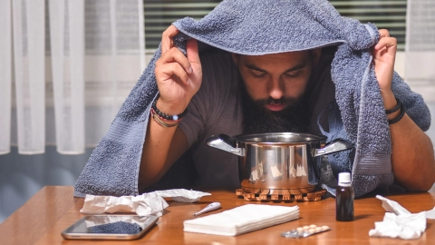 Ein Mann sitzt an einem Tisch, auf dem Taschentücher, Fieberthermometer, Tabletten, Nasentropfen und ein Topf stehen. Der Mann hält ein Handtuch über seinen Kopf und beugt sich über den Topf.