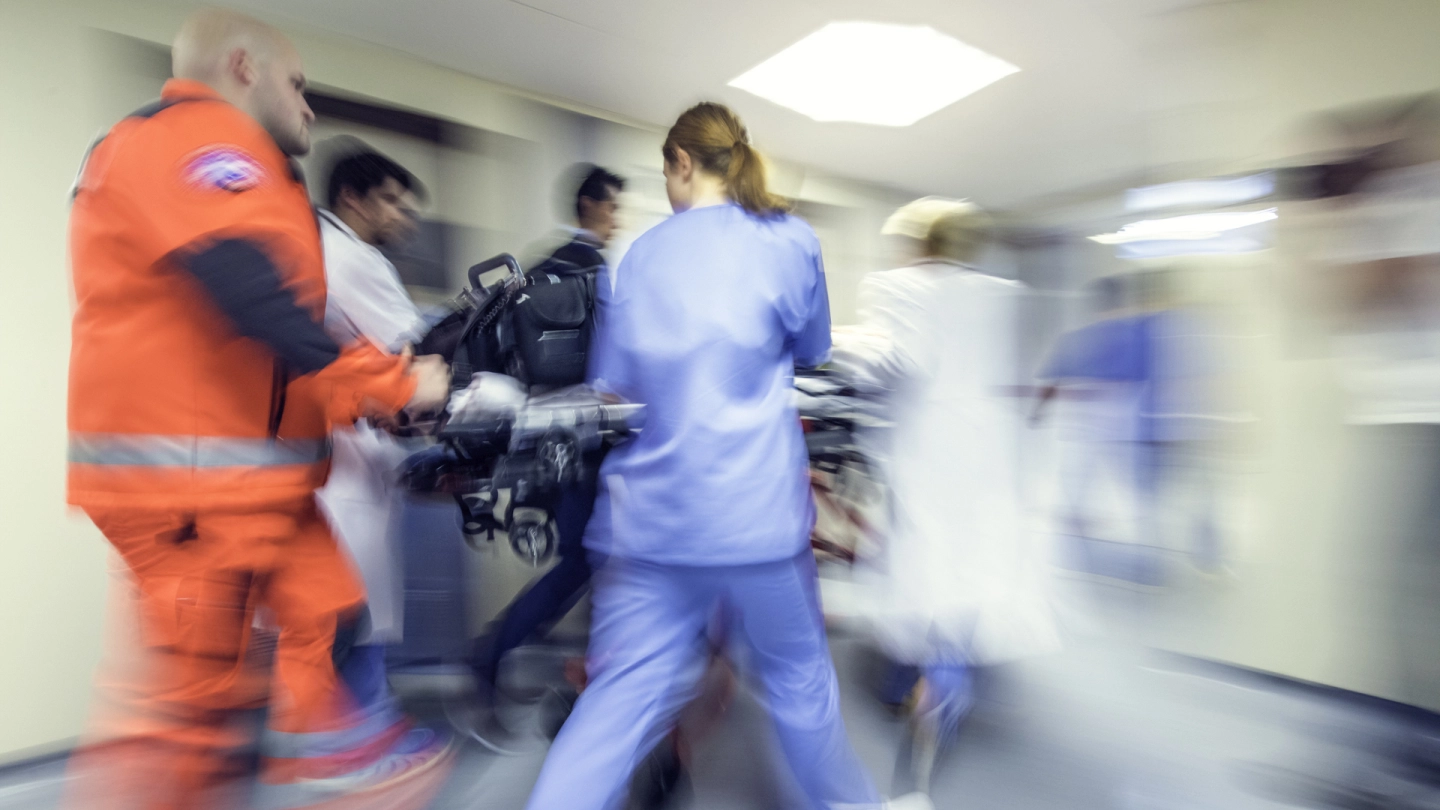 Ein Notfallsanitäter, eine Krankenschwester und ein Notarzt schieben eine Trage mit einem Patienten durch einen Flur im Krankenhaus.