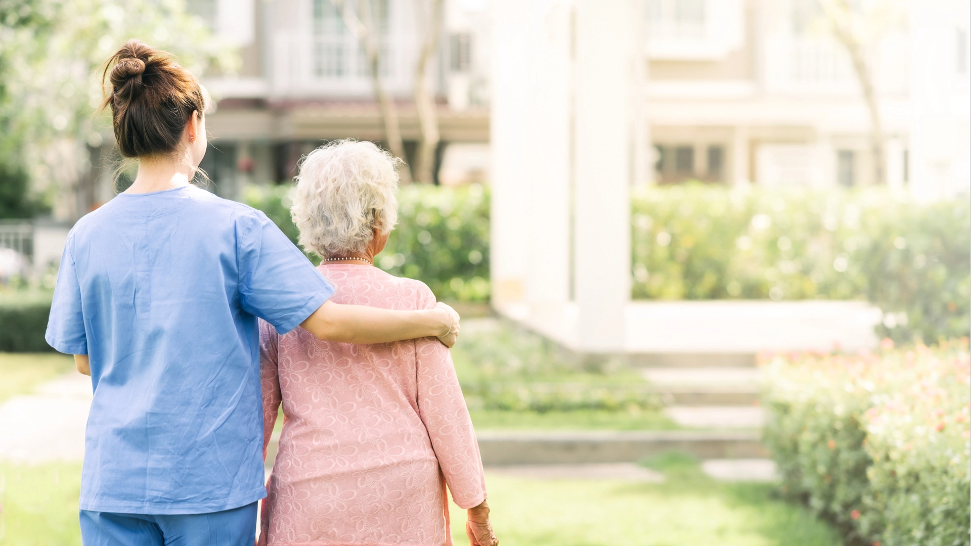Eine Pflegerin geht mit einer alten Dame in einer Grünanlage spazieren und legt den rechten Arm um ihre Schulter.