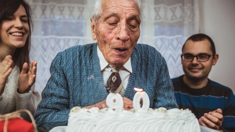 Prävention in Pflegeeinrichtungen: Ein festlich gekleideter älterer Herr pustet die Kerze einer Geburtstagstorte aus, die mit der Zahl 90 geschmückt ist. Zwei jüngere Menschen stehen um ihn herum, lächeln und applaudieren.
