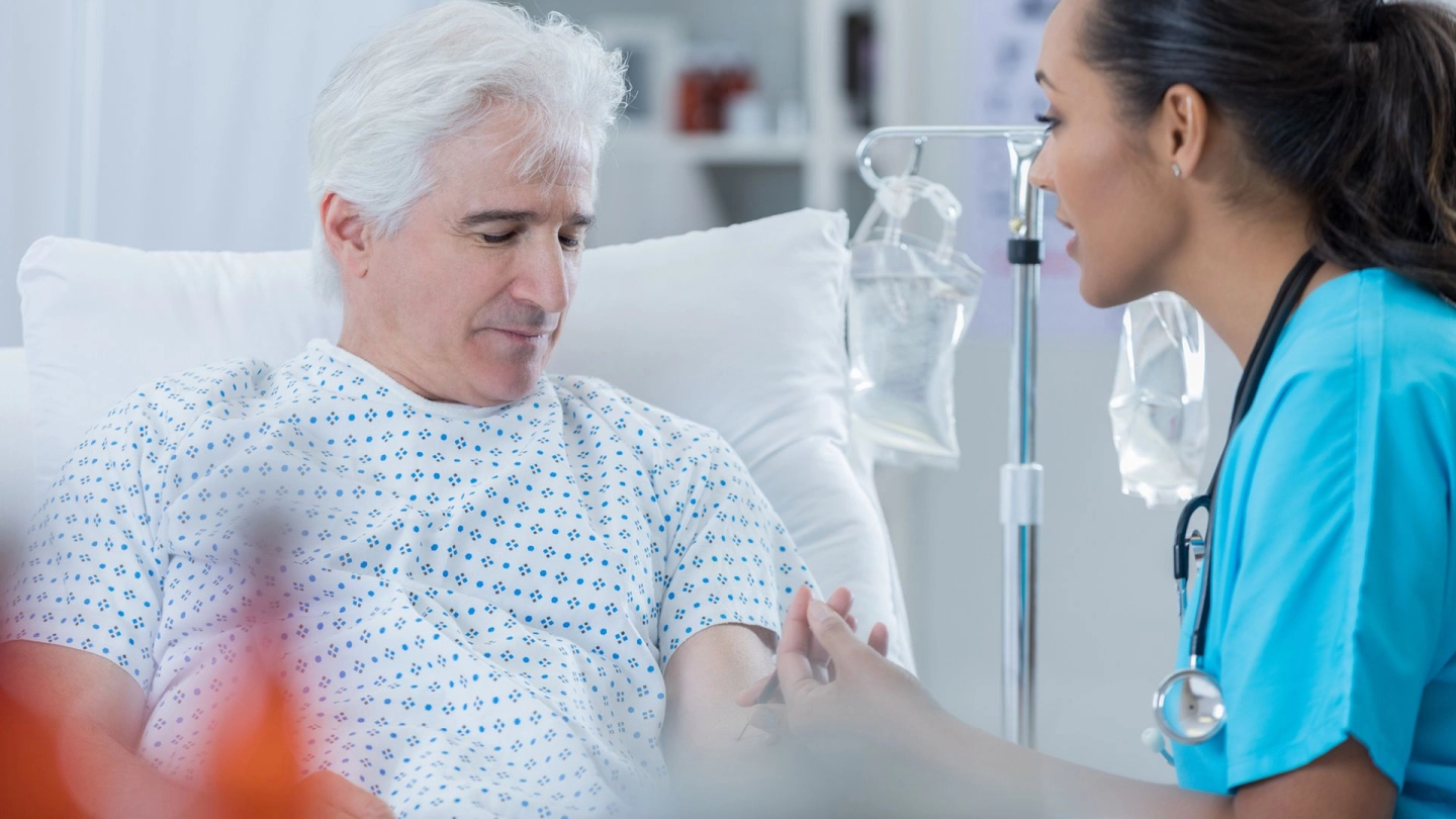 Prostatakrebs: Ein älterer Mann sitzt in einem Krankenhausbett. Er schaut nach unten, ihm gegenüber sitzt eine Krankenschwester. Sie schaut ihn an und entnimmt ihm Blut.