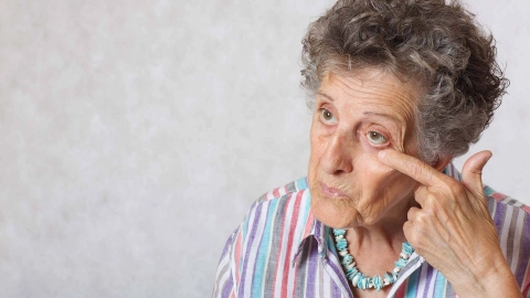 Eine ältere Dame fasst sich prüfend ans Auge, als würde sie sich im Spiegel betrachten.