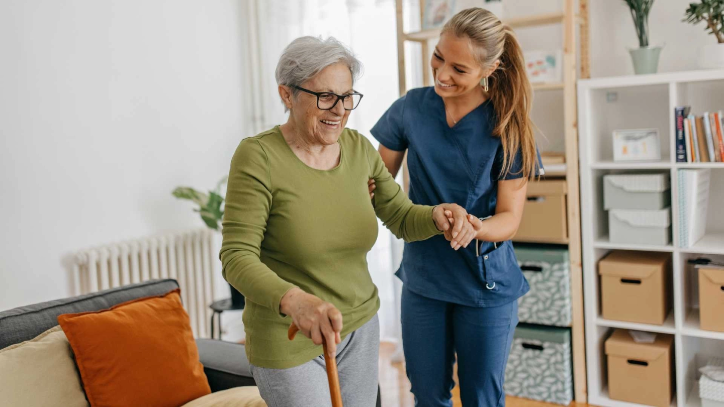 Медсестра поддерживает пожилую женщину. Пожилая женщина опирается на трость. Обе улыбаются.