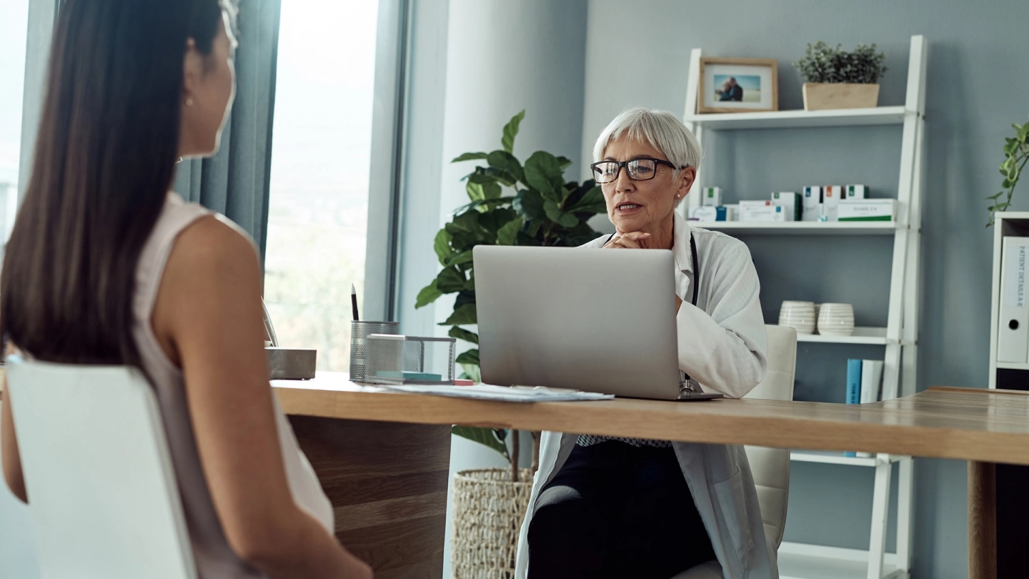 امرأة تجلس في غرفة العلاج. وهناك طبيبة تجلس أمامها وتنظر إلى الحاسب المحمول الخاص بها.