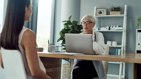 Eine Frau sitzt in einem Behandlungszimmer. Ihr gegenüber sitzt eine Ärztin und guckt auf ihren Laptop.