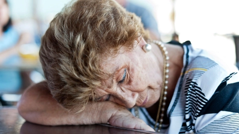 امرأة مسنة تسند رأسها على ذراعها التي تتكئ على منضدة. ويبدو عليها النعاس.