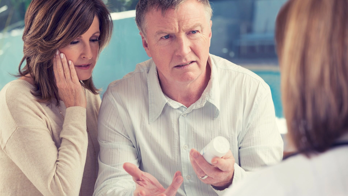 Ein Mann und eine Frau einer Ärztin gegenüber. In einer Hand hält der Mann ein Medikament. Er schaut die Ärztin fragend an.