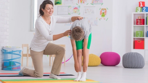 Улыбающаяся женщина-физиотерапевт поддерживает мальчика, который стоит, наклонившись вперед, и пытается дотянуться руками до своих стоп.