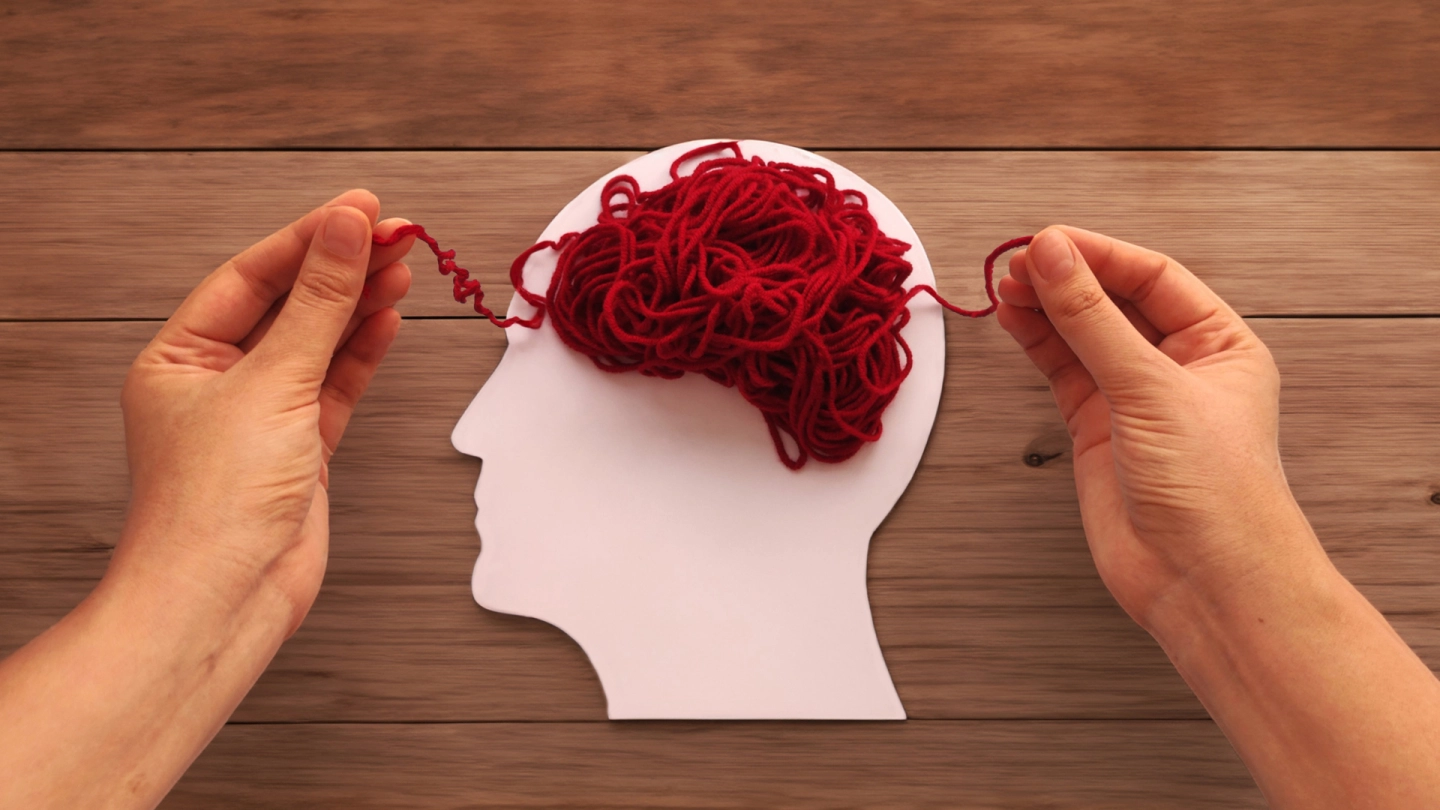 Ein rotes Wollknäuel liegt auf einer weißen Silhouette eines Kopfes. Von unten ragen zwei Hände ins Bild, die die losen Enden des Wollknäuels rechts und links des Kopfes festhalten.