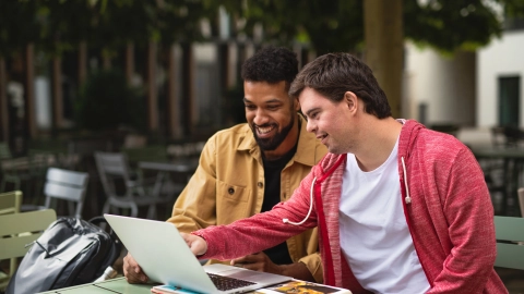 Ein junger Mann mit Behinderung und ein weiterer junger Mann schauen gemeinsam auf einen Laptop.