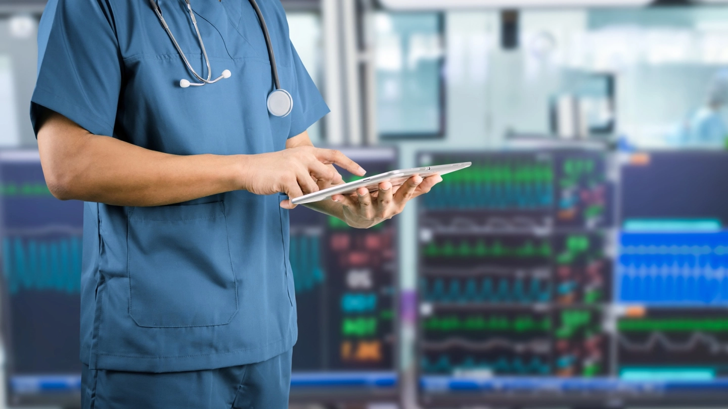 Eine medizinische Fachkraft bedient ein Tablet, im Hintergrund sind Monitore zur Kreislaufüberwachung sichtbar.