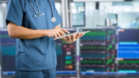Eine medizinische Fachkraft bedient ein Tablet, im Hintergrund sind Monitore zur Kreislaufüberwachung sichtbar.