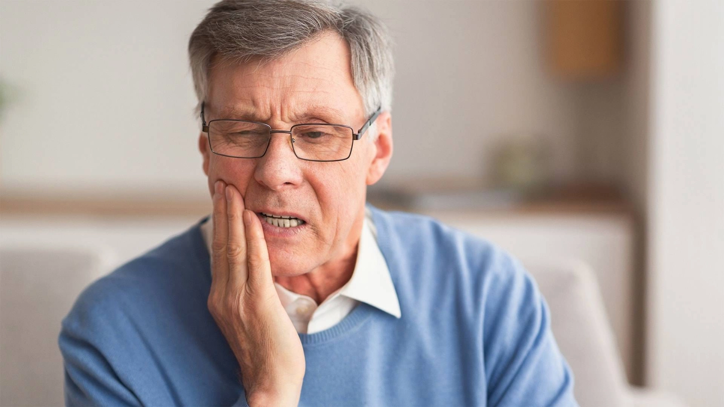 Ein gepflegter älterer Herr fasst sich besorgt an die rechte Wange; sein Gesicht ist schmerzverzerrt.