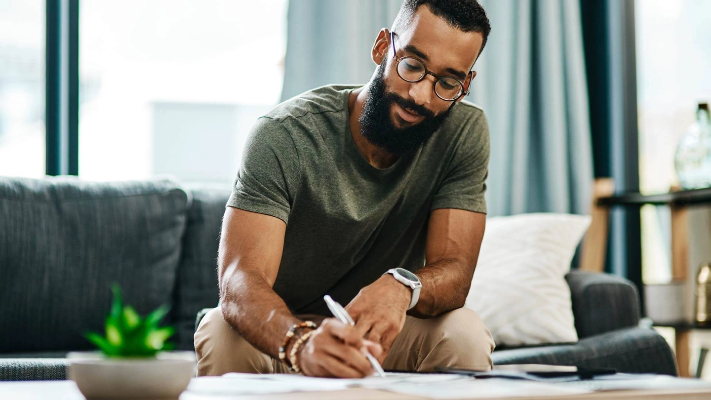 Ein Mann sitzt am Wohnzimmertisch. Er hält einen Stift in der Hand und füllt ein Formular aus.