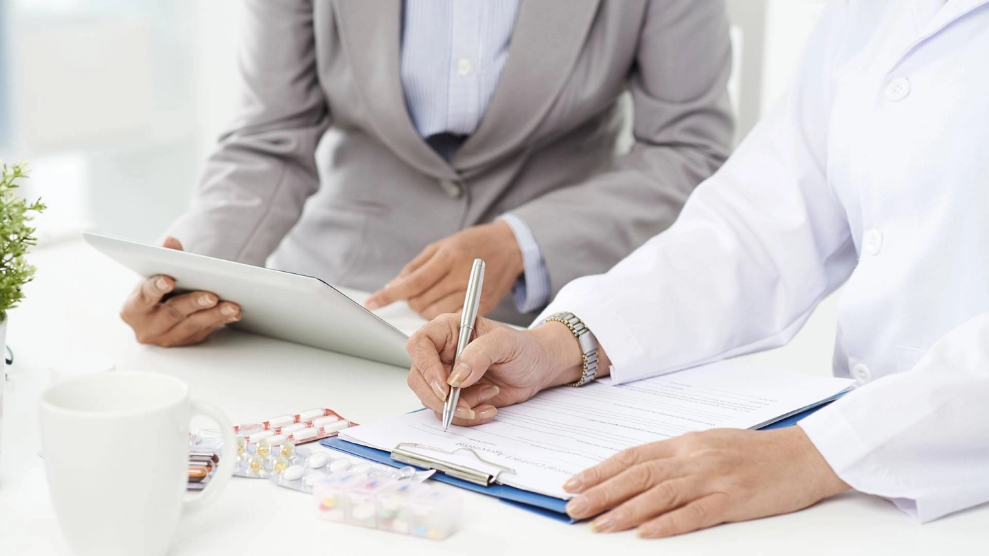 شخص يرتدي معطفًا أبيض ويملأ مستندًا. بجانبه يجلس شخص يشير إلى جهاز لوحي.