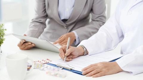 Eine Person im weißen Kittel füllt ein Dokument aus. Daneben sitzt eine Person, die auf ein Tablet zeigt.